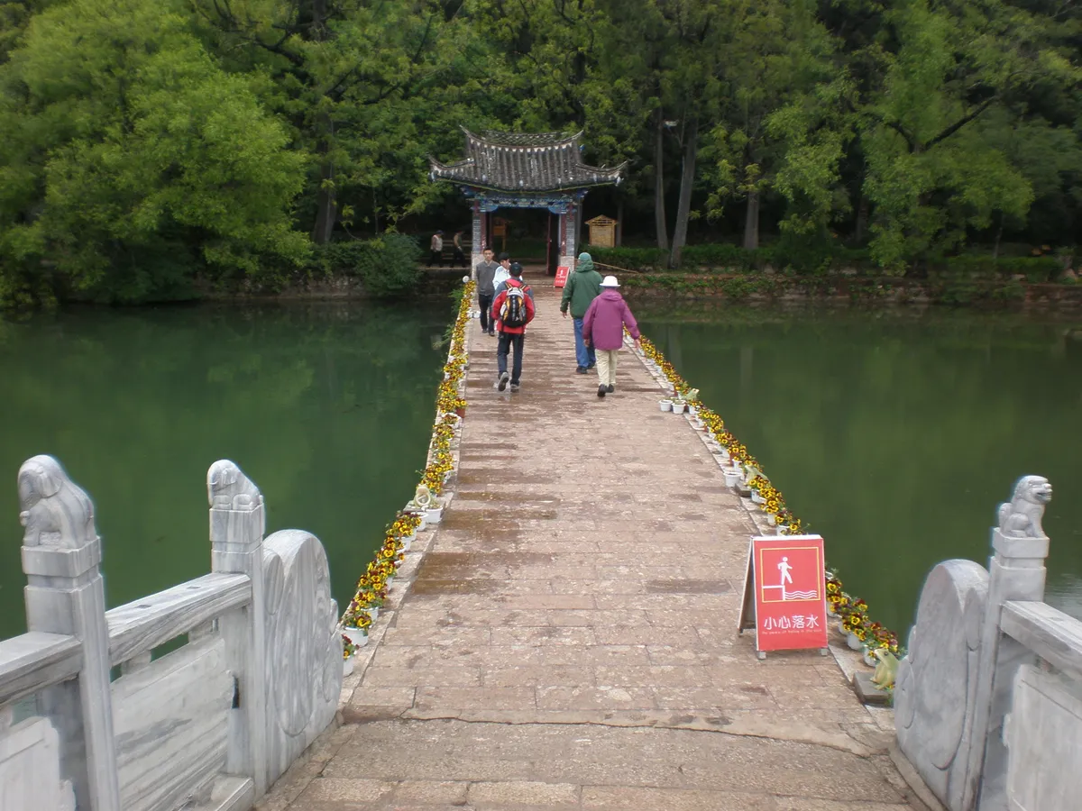 A stone footbridge across Black Dragon Pool leading to a traditional pavilion, framed by trees in Lijiang