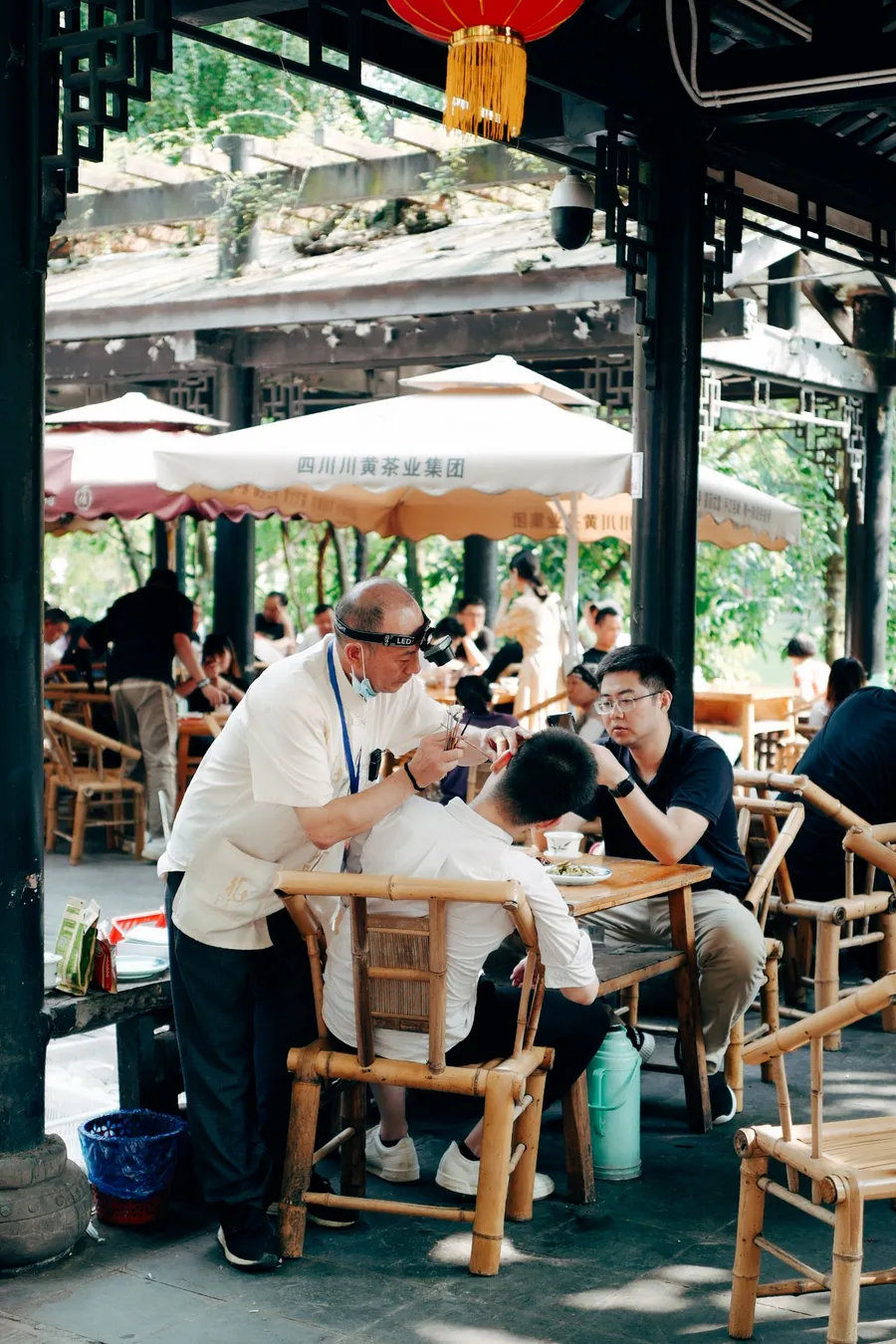 An ear cleaner at work on a seated customer under the umbrellas of an outdoor Sichuan teahouse, bamboo chairs and red lanterns around them