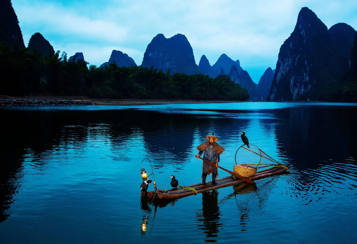 A cormorant fisherman standing on a bamboo raft with lantern and trained birds at dusk on the Li River, karst peaks behind