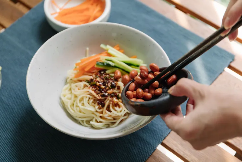 A bowl of noodles in chili oil with peanuts being added by hand
