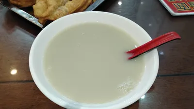 A bowl of grey-green douzhir with a red spoon, fried dough on the side, at a Beijing breakfast shop