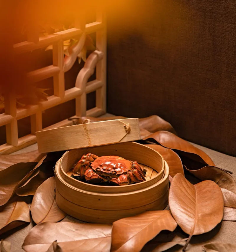A whole steamed hairy crab in a bamboo steamer basket surrounded by autumn leaves