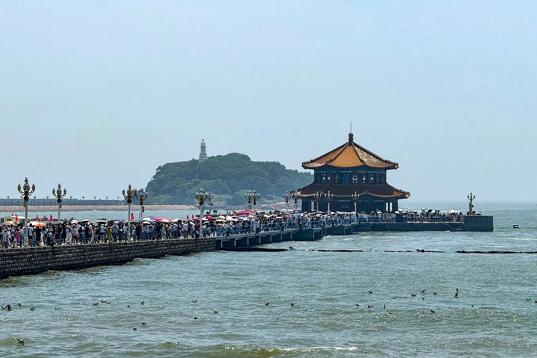Crowds walking Zhanqiao Pier toward its double-roofed pavilion, with Little Qingdao islet and lighthouse behind