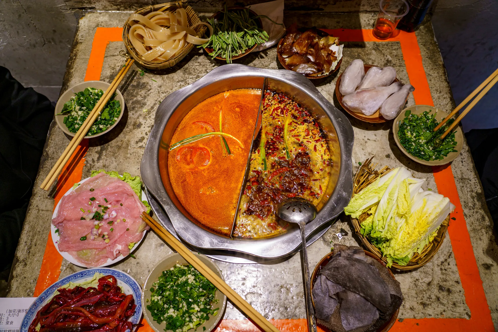 A split Sichuan hotpot with spicy and mild broths, surrounded by raw ingredients