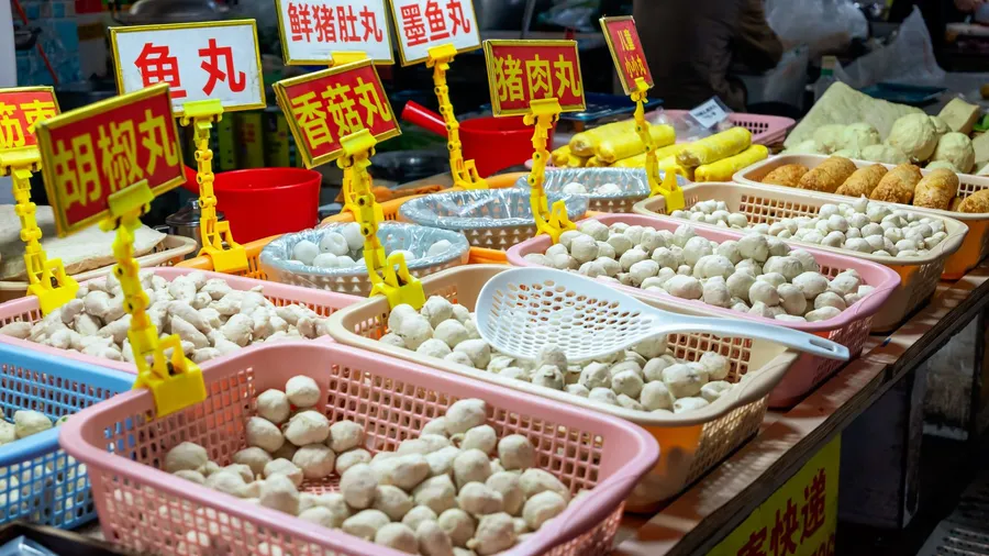 A Chinese market stall selling fish balls and meatballs from baskets, handwritten yellow price signs above each tray