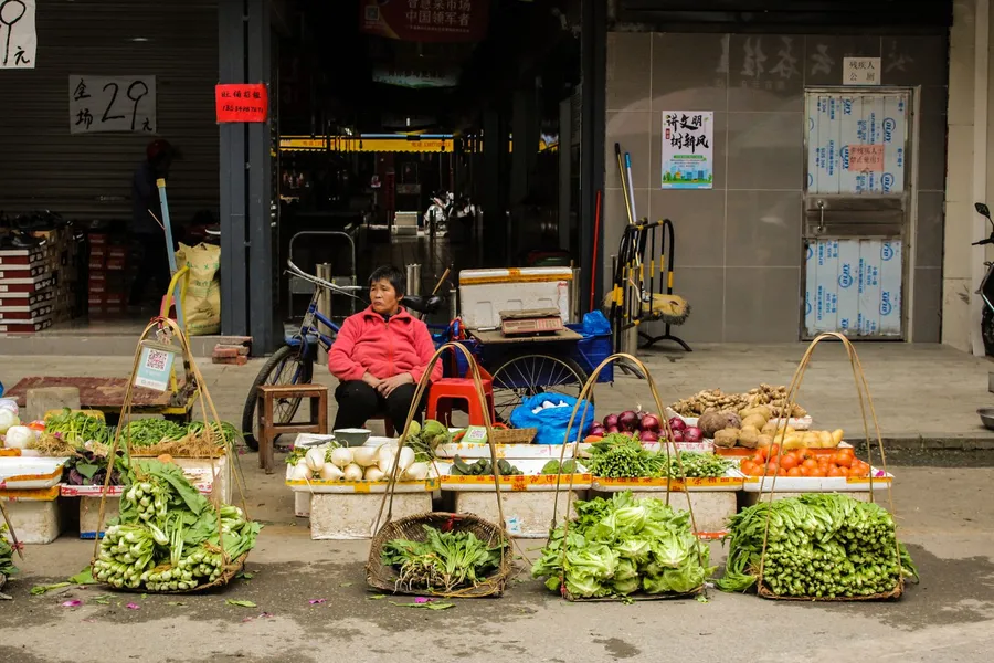 A street vegetable stall in China, produce laid out in baskets on the pavement with the vendor sitting behind