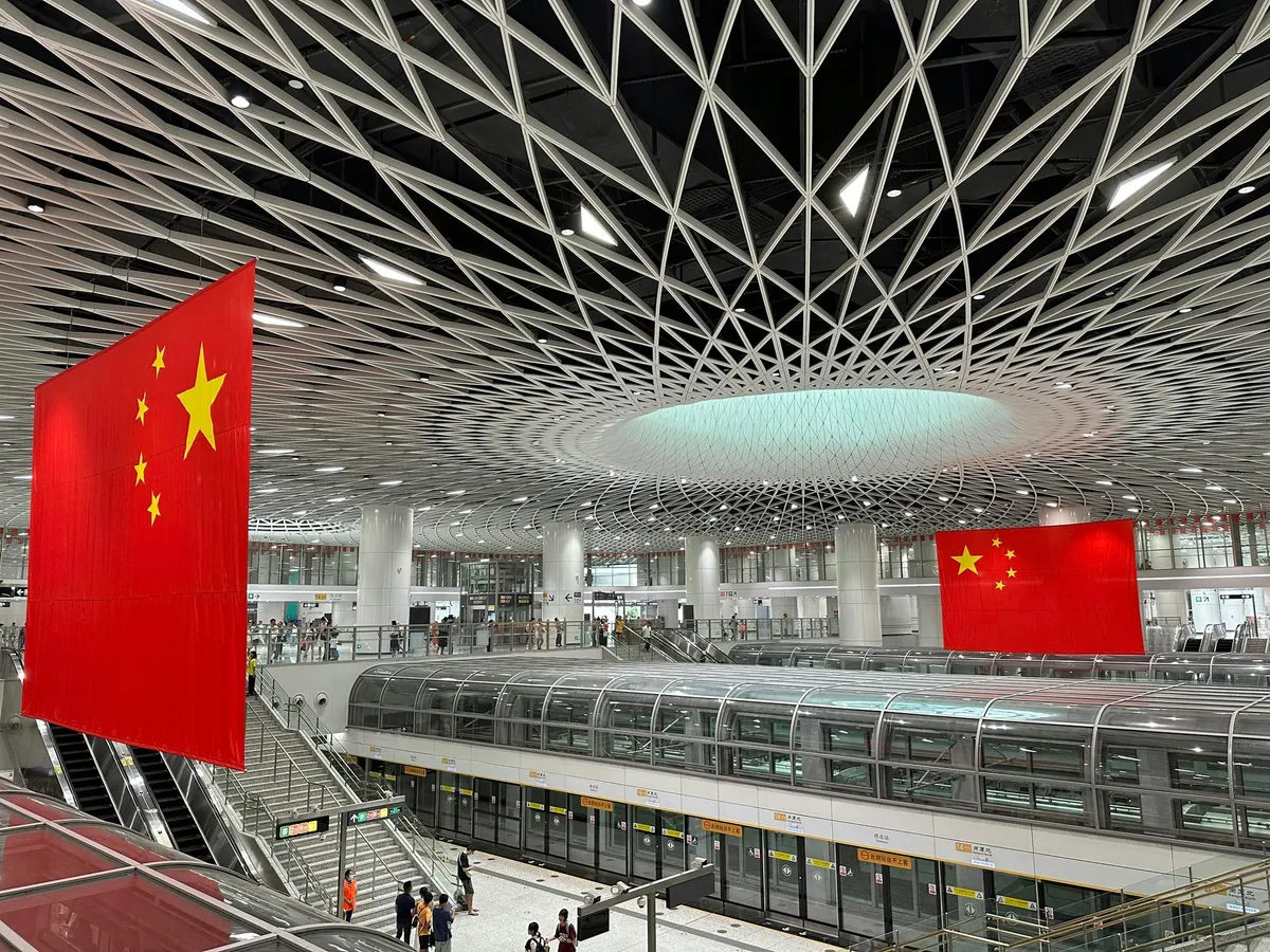 A large Chinese railway station concourse with national flags hanging above the platforms and escalators