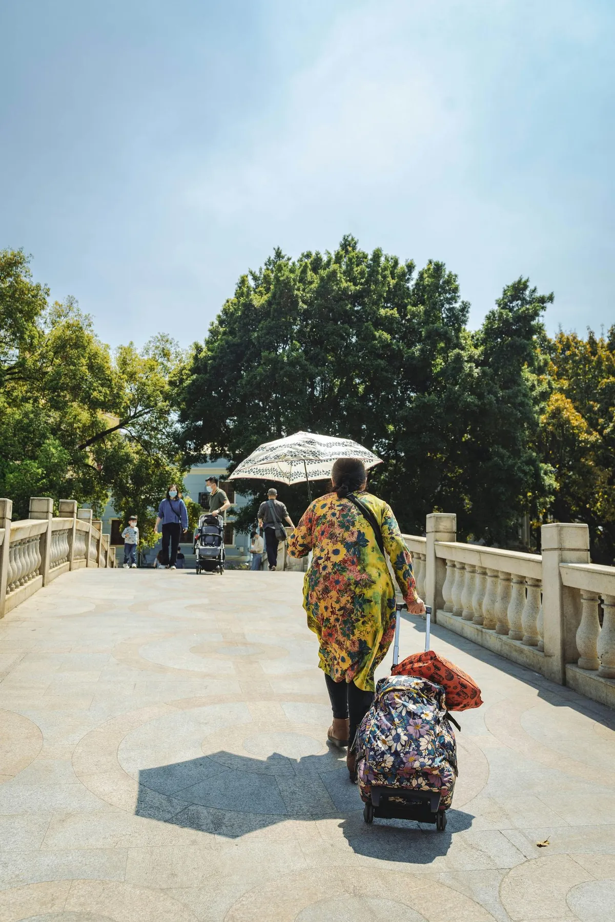 A traveler pulling a rolling suitcase while holding an umbrella for shade on a paved path in China