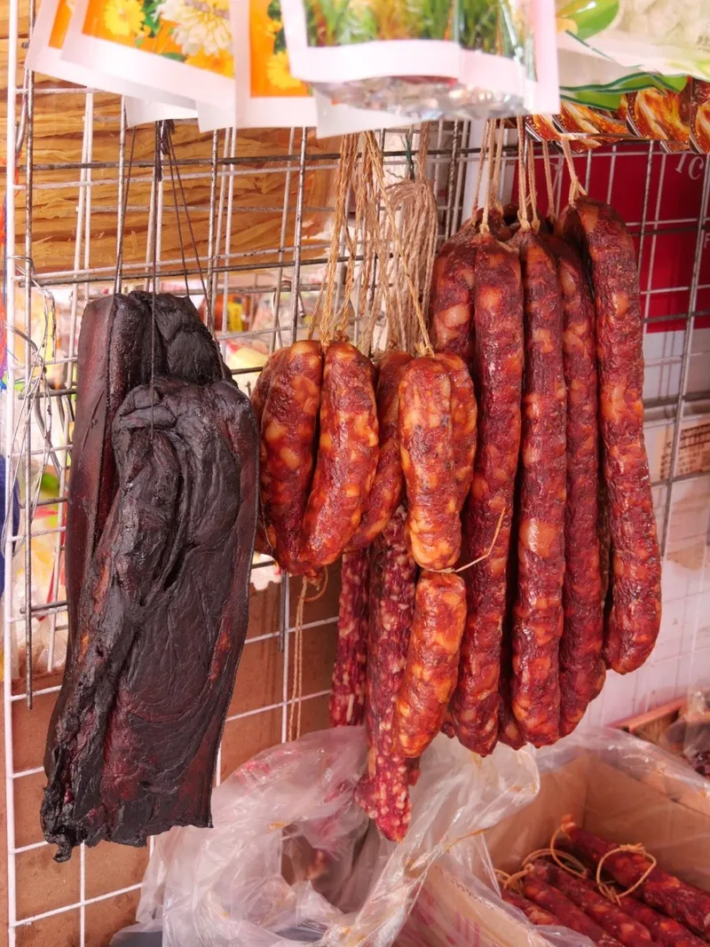 Dark, wood-smoked cured pork belly beside strings of Hunan-style sausages at a market stall